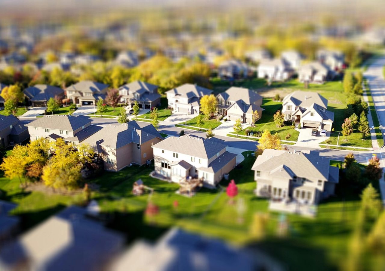 Tilt-shift aerial photo of a suburban neighborhood with green lawns and roads.